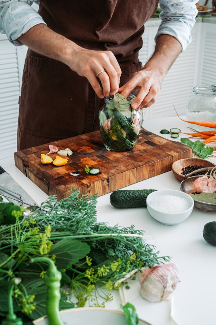 A man prepares fresh herbs and vegetables for pickling indoors on a wooden board.